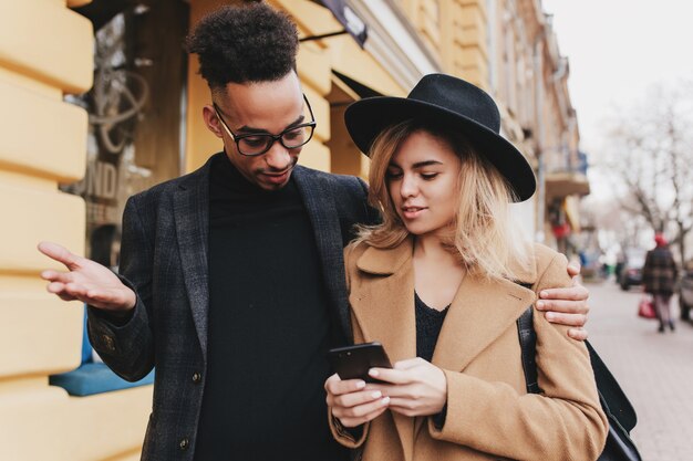 Surprised curly mulatto guy embracing magnificent blonde woman. Attractive fair-haired lady walking down the street and talking around with african male friend.