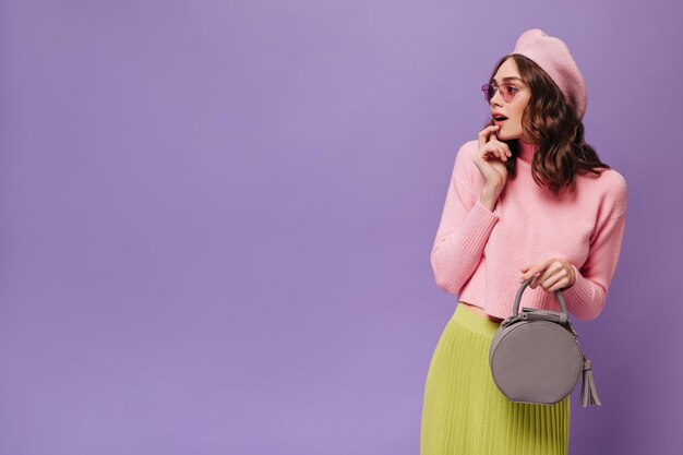 Surprised curly woman in beret looks to left Charming lady in pink sweater sunglasses and green skirt holds grey handbag