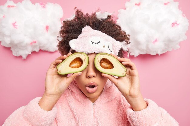 Surprised curly woman covers eyes with halves of avocado uses for natural cosmetic cares about skin keeps mouth opened from wonder wears sleepmask and pajama