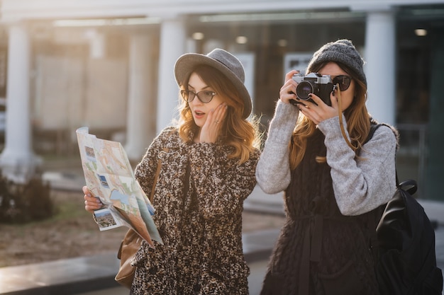 Удивленная кудрявая женщина в очках, глядя на карту, трогая лицо, пока ее друг фотографирует достопримечательности. Привлекательная женщина-путешественница гуляет с камерой и ее сестра ищет интересные места.