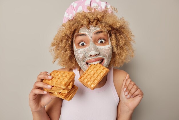 Surprised curly woman wears bathhat and casual t shirt bites waffle applies beauty mask on face for skin treatment looks wondered isolated over grey background Sweet food and wellness concept