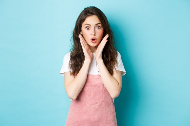 Surprised cute girl gasping amazed, saying wow and looking at camera astonished, hear interesting news, checking out promo offer, standing against blue background