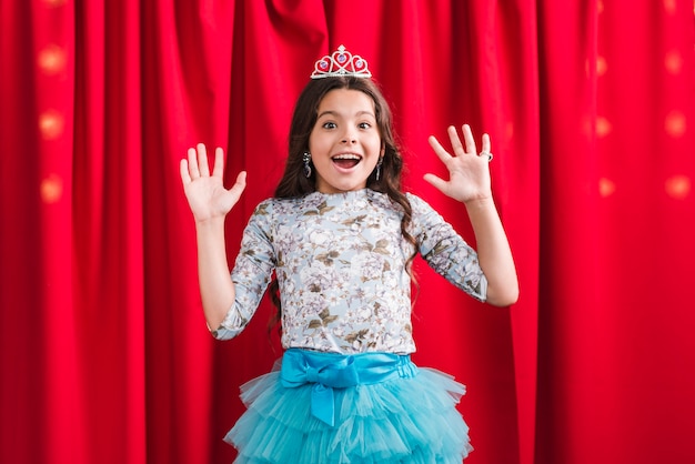 Surprised cute girl wearing crown standing in front of red curtain