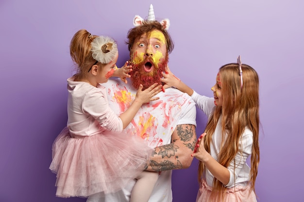 Surprised dad and two girls play together at home, paint faces with watercolours, have fun, show hands painted in bright colours, isolated over purple wall. Family portrait. Fatherhood