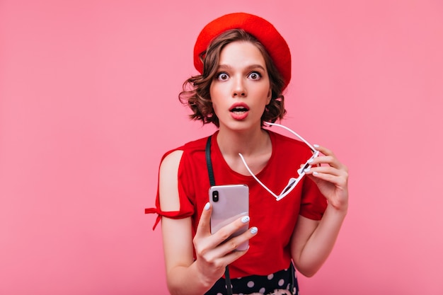 Surprised dark-haired girl posing with phone. Well-dressed young lady in beret expressing amazement.
