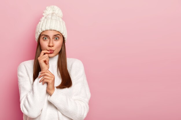 Surprised dark haired woman looks surprisingly, keeps lips rounded, wears snow white winter hat and sweater, dressed in warm outfit poses against pink wall