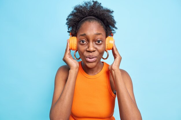 Surprised dark skinned young woman keeps hands on stereo headphones