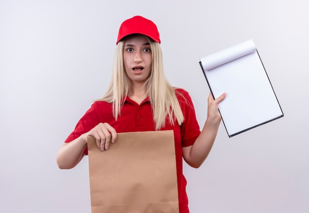 Surprised delivery young girl wearing red t-shirt and cap in dental brace holding paper pocket and showing clipboard on her hand on isolated white background