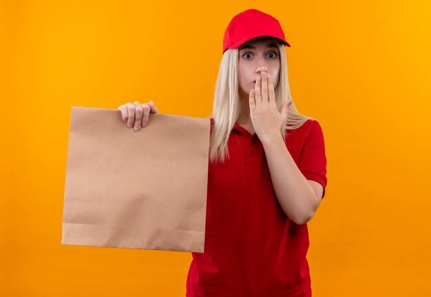孤立したオレンジ色の背景に手で赤いTシャツと紙のポケットで覆われた口を保持しているキャップを身に着けている驚きの配達の少女