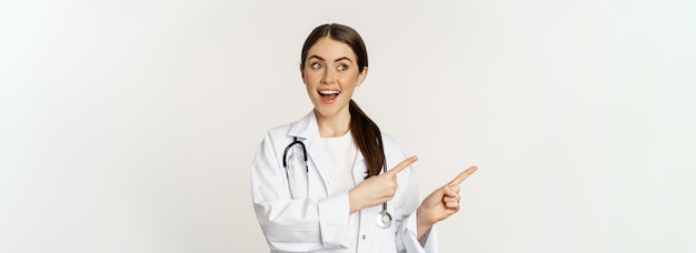 Бесплатное фото Удивленный врач показывает пальцем влево и смотрит на баннер с логотипом, показывающий рекламу клиники, стоящую на белом фоне