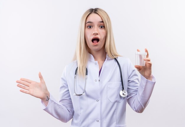 Surprised doctor young blonde girl wearing stethoscope and medical gown holding empty can raised hand on isolated white background