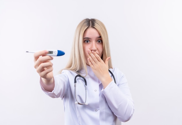 Foto gratuita stetoscopio da portare sorpreso della giovane ragazza bionda del medico e termometro della tenuta dell'abito medico fuori alla bocca coperta della macchina fotografica su fondo bianco isolato