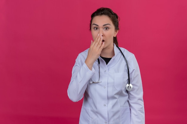 Free photo surprised doctor young girl wearing medical gown and stethoscope covered mouth on isoleted red background