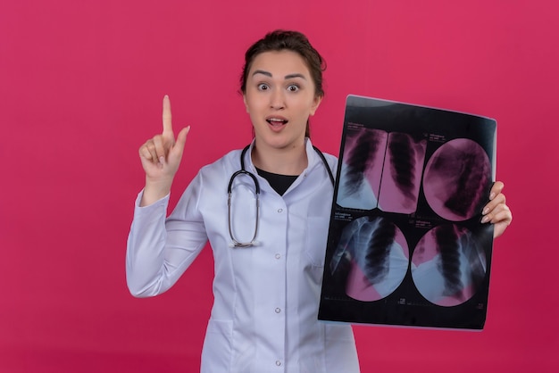 Удивленная молодая девушка-врач в медицинском халате и стетоскопе держит рентгеновский снимок и указывает вверх на изолированном красном фоне