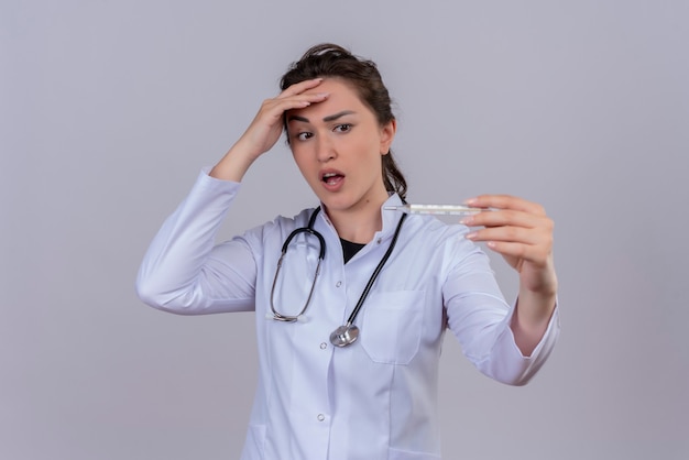 Surprised doctor young girl wearing medical gown wearing stethoscope holding thermometer and put her hand on forehead on white background