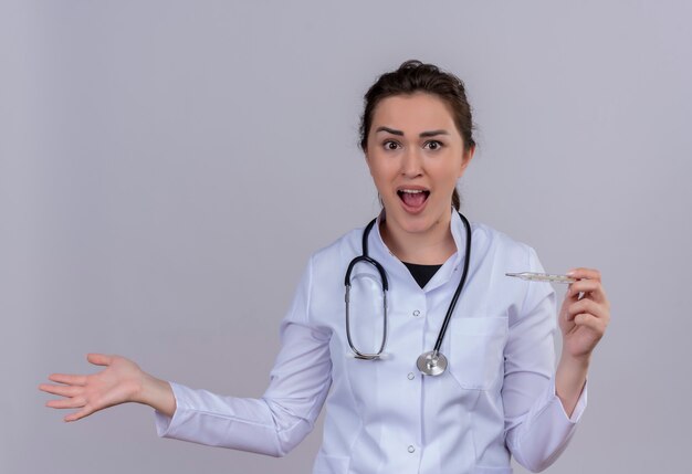 Surprised doctor young girl wearing medical gown wearing stethoscope holding thermometer and spread hand on white background