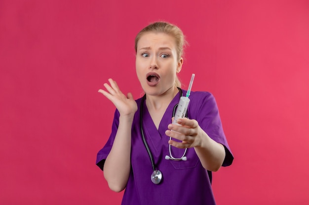 Surprised doctor young girl wearing purple medical gown and stethoscope holding syringe on isolated pink background