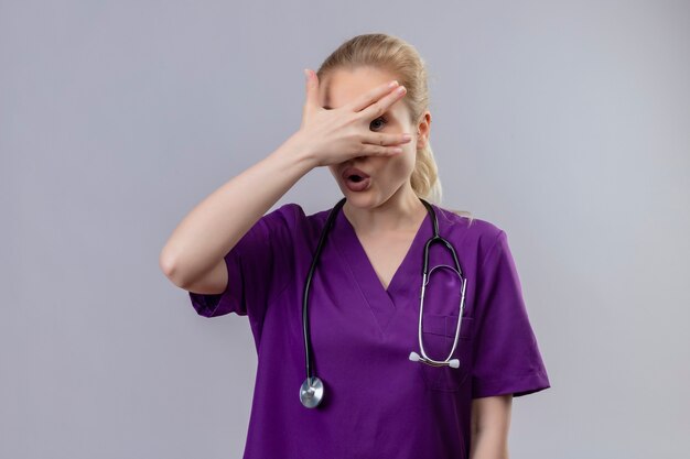 Удивленная молодая девушка-врач в фиолетовом медицинском халате и стетоскопе положила руку на глаза на изолированном белом фоне