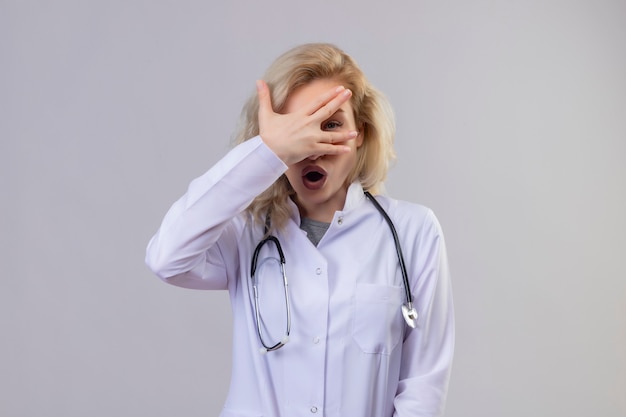 Удивленная молодая девушка-врач со стетоскопом в медицинском халате закрыла глаза на белом фоне