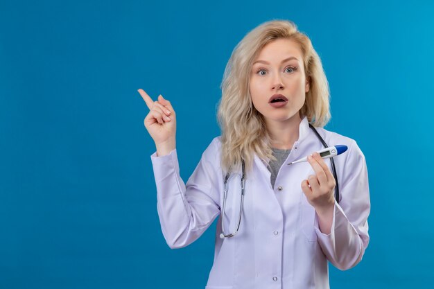 Surprised doctor young girl wearing stethoscope in medical gown holding thermometer points to side on blue background