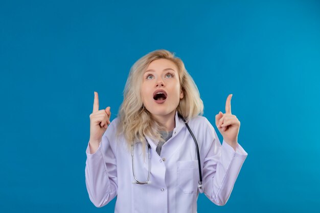 Surprised doctor young girl wearing stethoscope in medical gown points to up on blue background