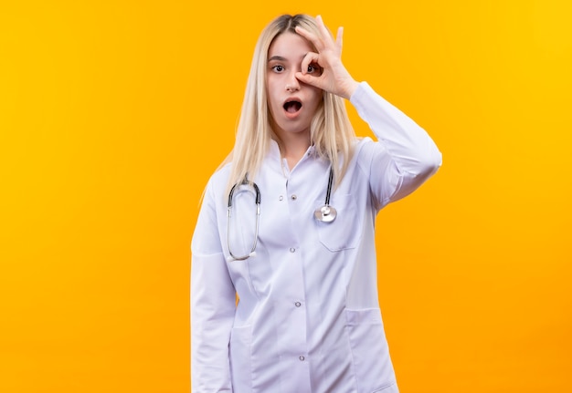 Surprised doctor young girl wearing stethoscope in medical gown showing look gesture on isolated yellow background