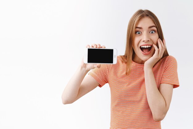 Surprised, eager and enthusiastic good-looking young caucasian woman in striped t-shirt, holding smartphone horizontally, showing black mobile display and touch cheek from amazement and joy