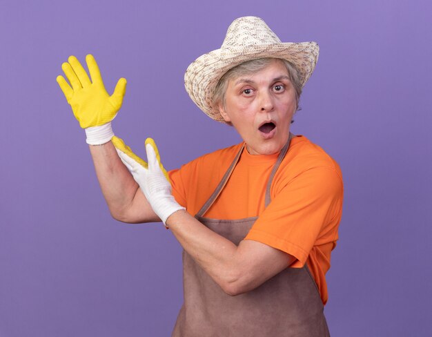 Удивленная пожилая женщина-садовник в садовой шляпе и перчатках, указывая в сторону двумя руками на фиолетовом