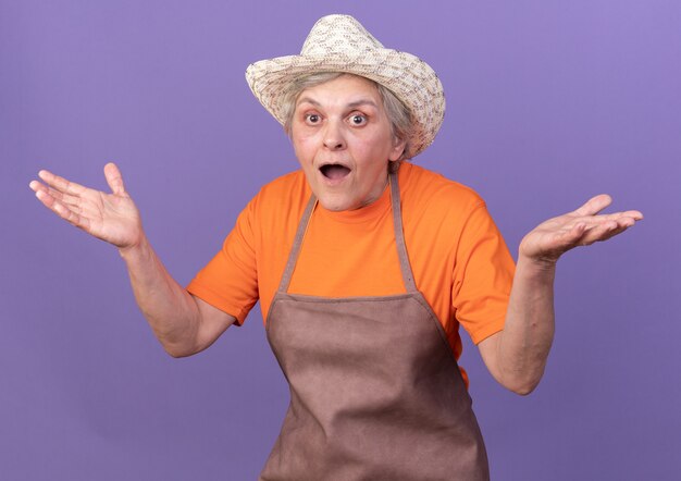 Free photo surprised elderly female gardener wearing gardening hat holds hands open on purple