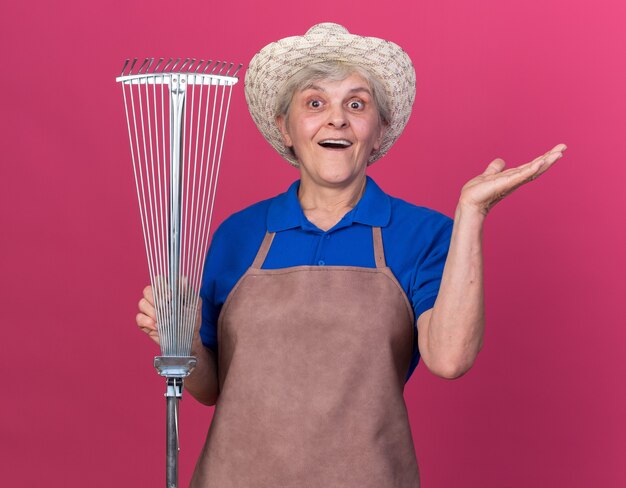 Surprised elderly female gardener wearing gardening hat holds leaf rake and keeps hand open