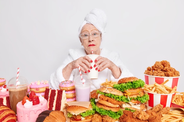 Surprised elderly lady with red lipstick has imbalancd nutrition eats different tasty junk food drinks cocktail containing much sugar dressed in domestic clothes