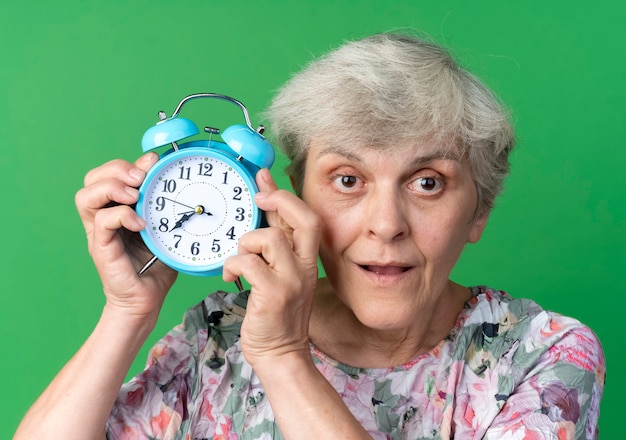 Удивленная пожилая женщина держит будильник рядом с головой, изолированную на зеленой стене