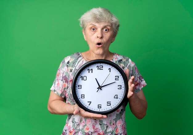 Удивленная пожилая женщина держит часы, изолированные на зеленой стене