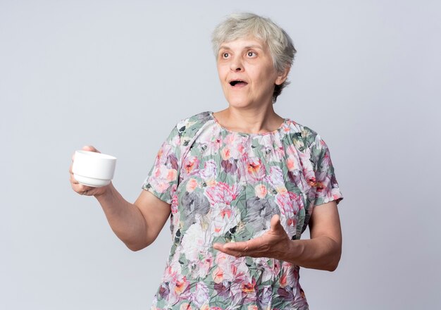 Surprised elderly woman holds cup looking and pointing at side isolated on white wall