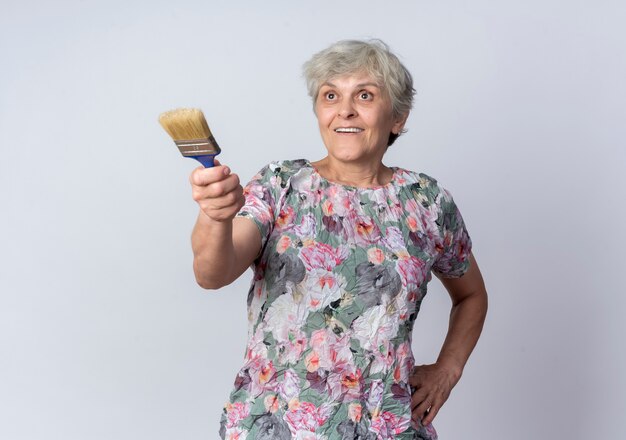 Surprised elderly woman holds and points forward with paint brush isolated on white wall