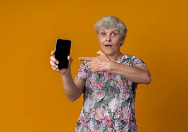 Surprised elderly woman holds and points at phone isolated on orange wall