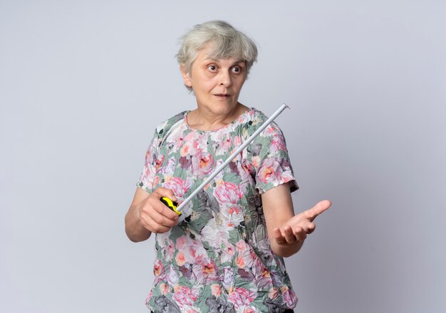 Удивленная пожилая женщина держит рулетку, глядя и указывая вперед, изолированную на белой стене