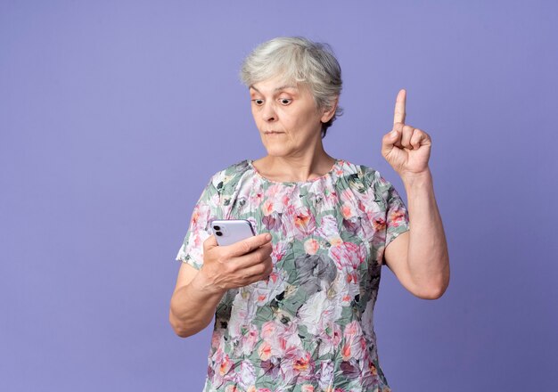 Удивленная пожилая женщина смотрит на телефон и указывает вверх, изолированную на фиолетовой стене