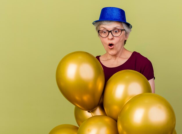 Surprised elderly woman in optical glasses wearing party hat holds and looks at helium balloons isolated on olive green wall with copy space