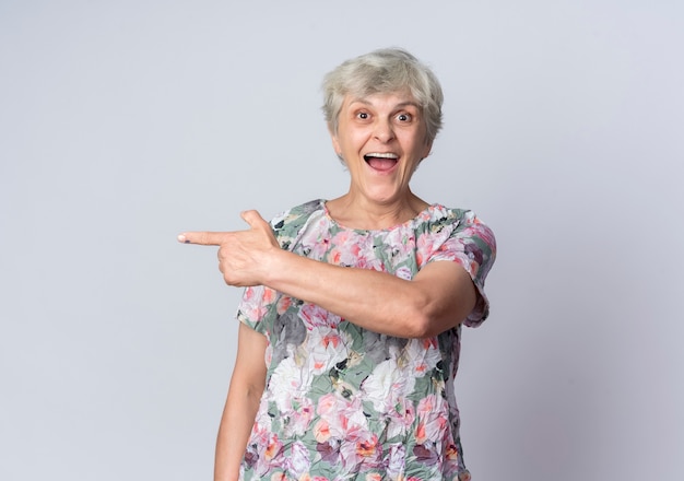 Surprised elderly woman points at side looking isolated on white wall