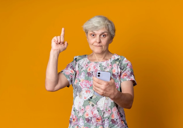 Surprised elderly woman points up holding phone isolated on orange wall