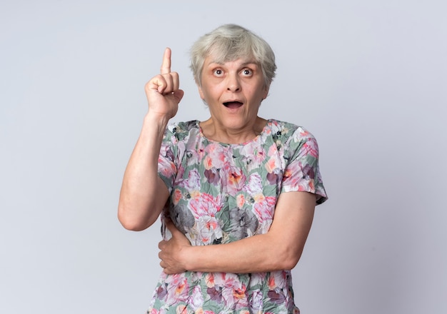 Surprised elderly woman points up looking forward isolated on white wall