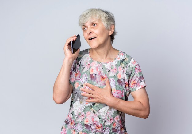 Surprised elderly woman puts hand on chin holds phone looking at side isolated on white wall