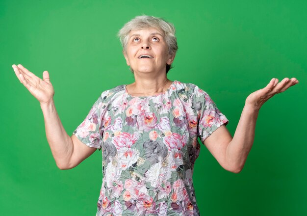 Surprised elderly woman raises hands looking up isolated on green wall