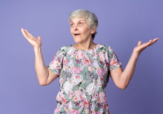 Surprised elderly woman stands with open hands looking at side isolated on purple wall
