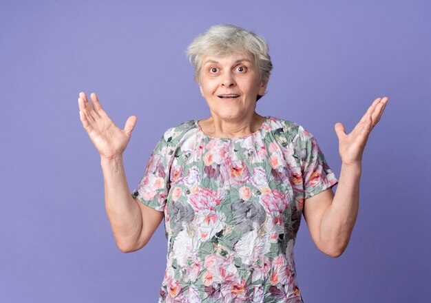 Surprised elderly woman stands with raised hands isolated on purple wall