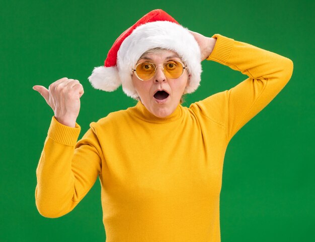 Surprised elderly woman in sun glasses with santa hat pointing at side 