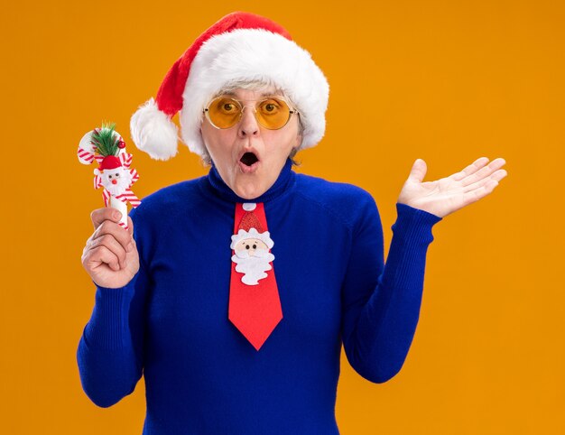 Surprised elderly woman in sun glasses with santa hat and santa tie holding candy cane and keeping hand open isolated on orange background with copy space