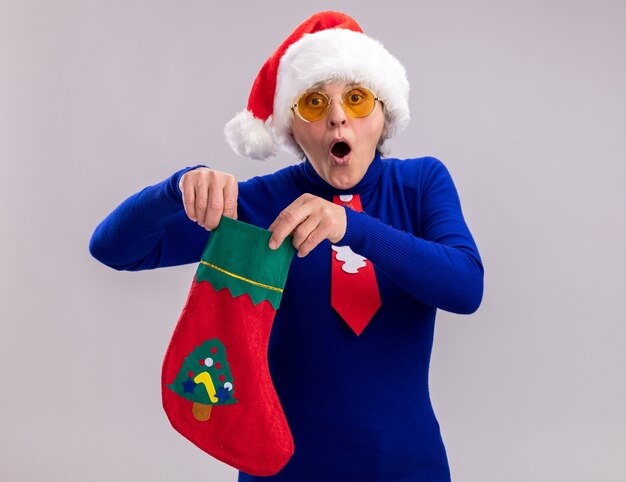 Surprised elderly woman in sun glasses with santa hat and santa tie holding christmas stocking isolated on white wall with copy space