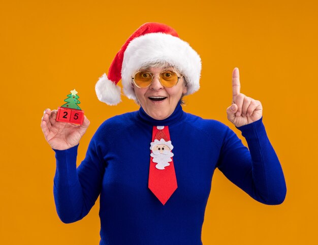 Surprised elderly woman in sun glasses with santa hat and santa tie holding christmas tree ornament and pointing up isolated on orange background with copy space